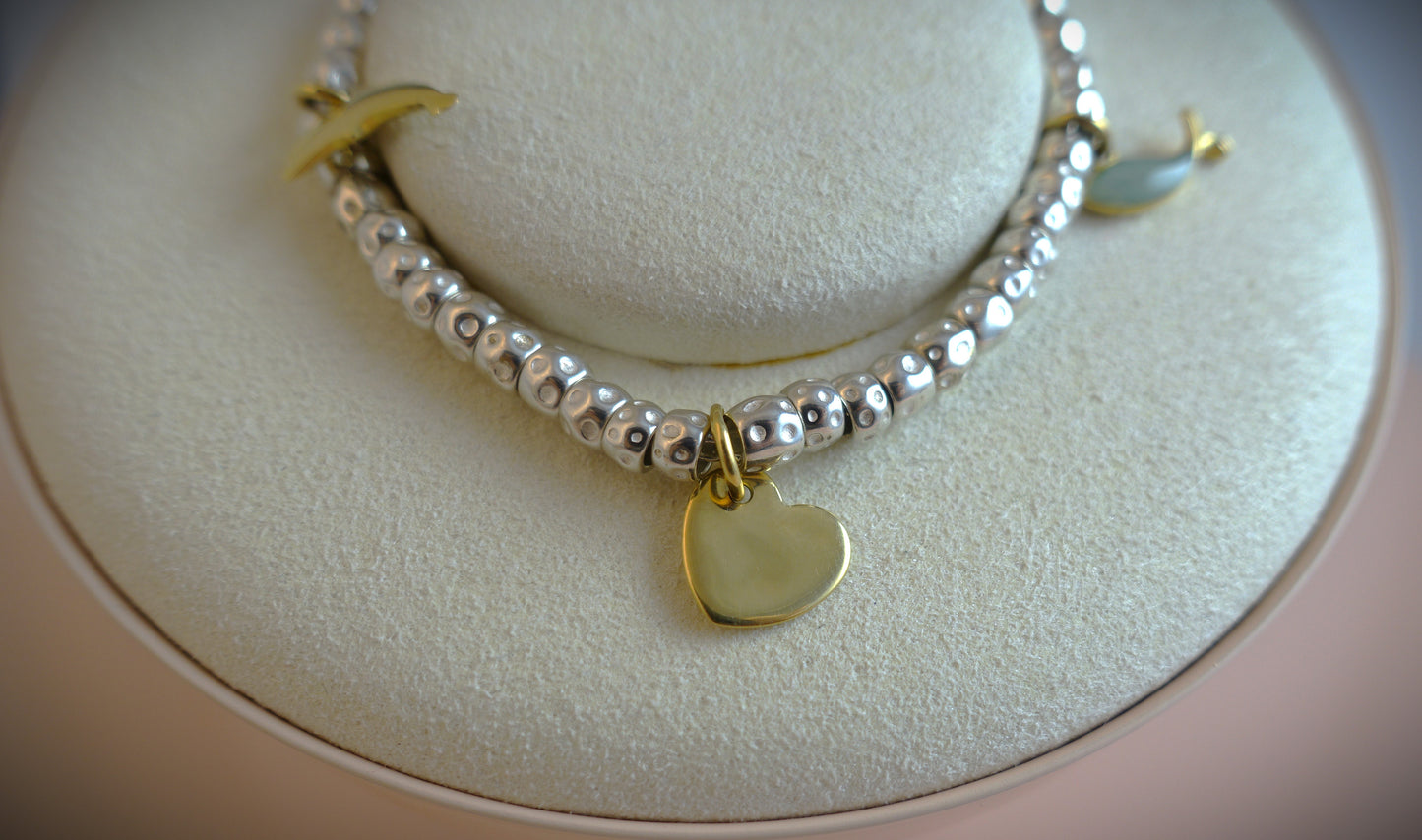 Das Dodo Armband Granelli von Ninas Schmuckhandel besteht aus einem Perlenarmband auf einem runden cremefarbenen Ständer mit 750 Gelbgold-Anhängern und Silberperlen, darunter ein Herzanhänger in der Mitte und eine Mondsichel aus 925 Sterling Silber.